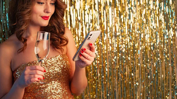Giovane donna beve champagne, tenendo uno smartphonee, divertirsi su uno sfondo oro lucido — Foto Stock
