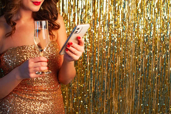 Giovane donna beve champagne, tenendo uno smartphonee, divertirsi su uno sfondo oro lucido — Foto Stock