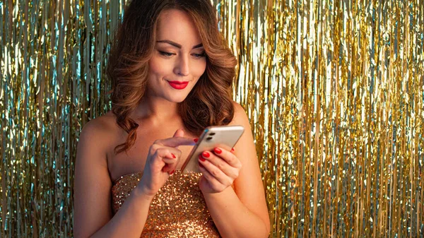 Young woman using phone on a shiny gold background — Stock Photo, Image