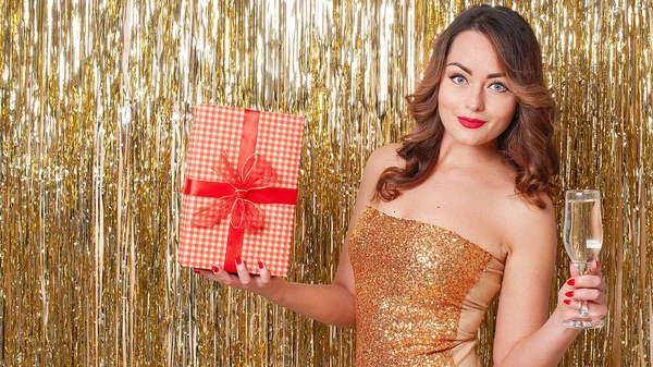 Giovane donna in possesso di una scatola regalo e beve champagne su uno sfondo oro lucido — Foto Stock