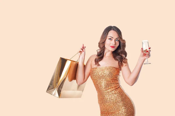 Женщина в модной одежде с золотым бумажным пакетом и бокалом игристого вина на бежевом фоне — стоковое фото