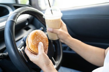 Asyalı kadın sürücü elinde buzlu kahve fincanı ve arabada yemek ve içmek için sandviç ekmeği tutuyor. Tehlikeli ve kaza riski var..