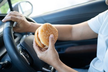 Asyalı kadın sürücü elinde buzlu kahve fincanı ve arabada yemek ve içmek için sandviç ekmeği tutuyor. Tehlikeli ve kaza riski var..