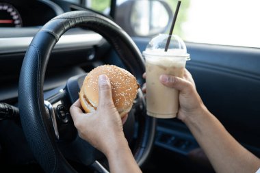 Asyalı kadın sürücü elinde buzlu kahve fincanı ve arabada yemek ve içmek için sandviç ekmeği tutuyor. Tehlikeli ve kaza riski var..