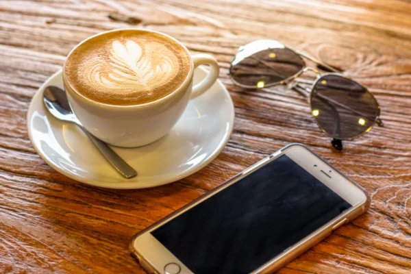 Taza de café arte latte con cuchara y plato en la corteza marrón hermoso fondo textura con luz cálida decorada con gafas de sol y teléfono móvil — Foto de Stock
