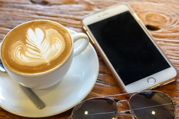 Taza de café arte latte con cuchara y plato en la corteza marrón hermoso fondo textura con luz cálida decorada con gafas de sol y teléfono móvil — Foto de Stock