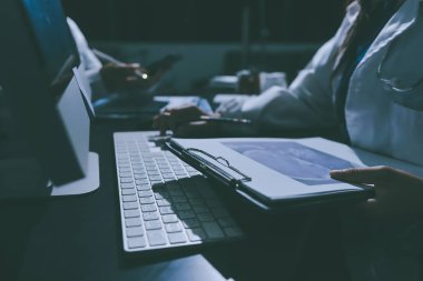 Two doctors and a female nurse meet at a table in the hospital, collaborating on medical tasks using laptops and computers