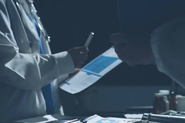 Two doctors and a female nurse meet at a table in the hospital, collaborating on medical tasks using laptops and computers