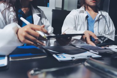 Two doctors and a female nurse meet at a table in the hospital, collaborating on medical tasks using laptops and computers