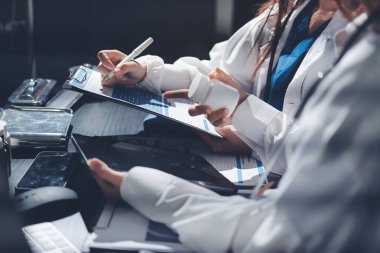 Two doctors and a female nurse meet at a table in the hospital, collaborating on medical tasks using laptops and computers