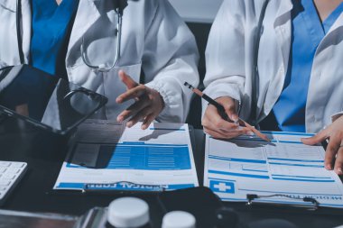 Two doctors and a female nurse meet at a table in the hospital, collaborating on medical tasks using laptops and computers