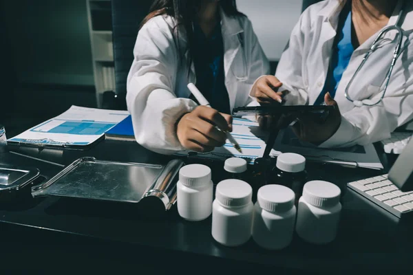 Two doctors and a female nurse meet at a table in the hospital, collaborating on medical tasks using laptops and computers