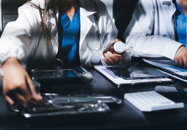 Two doctors and a female nurse meet at a table in the hospital, collaborating on medical tasks using laptops and computers