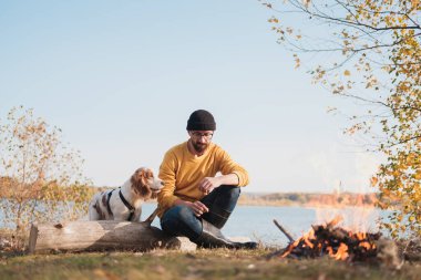 Adam gölün kenarında oturan köpeğine sarılıyor. Evcil hayvanlarla aktif dinlenme, maceralara çıkma, arkadaşlık: spanyel köpek ve sahibi açık havada oturur.