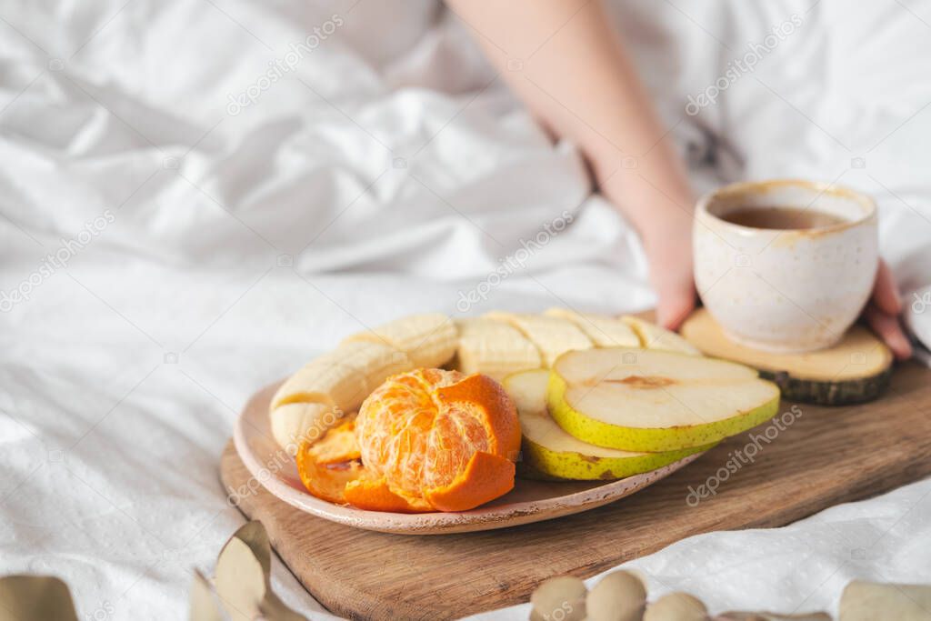 Having a breakfast in bed from a tray with fruits and tea in white clean bed. Mindful and quiet living, slow life, simple pleasures at home