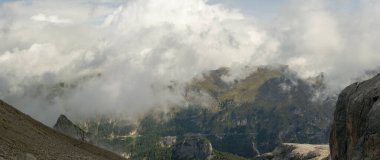 Alçak bulutlar, Dolomitler 'in dağlarındaki kayalık tepelerle çevrili ormanlık dağların üzerinde kıvrılır. Güneş ışığı sisi delip geçer, yeşil yamaçları aydınlatır, dolambaçlı dağ yolu ve kuzey İtalya 'da dramatik yüksek irtifa manzarası.