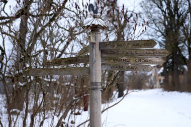 Kırsal bir ahşap levha, çıplak ağaçlar, yapraksız çalılar ve karları eşeleyen kuru otlarla çevrili, karlı bir kırsal bölgede duruyor. Sahne, karar verme mekanizmasını çağrıştırıyor..