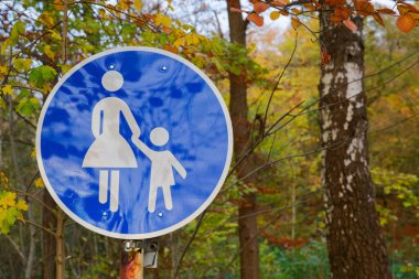 Ormanın kenarında mavi yuvarlak bir yaya bölgesi tabelası var. Açık figür ve parlak alan güvenli bir yürüyüş alanını işaret ederken, yüksek ağaçlar ormanda yumuşak bir yol çiziyor..