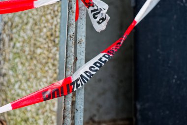 Kırmızı beyaz bant bir binanın köşesinde, direkler ve çitlerle çevrili. Sınırı işaretle, memurlar güvenlik ve kanıt yollarını kapatıyor. Komşular Almanya 'da belgelenirken geride duruyorlar.