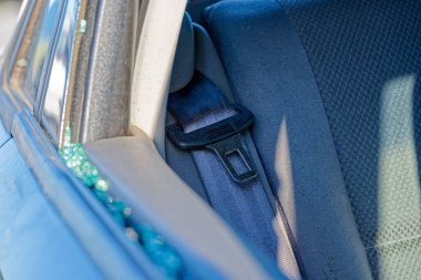 Bir araç yan camı parçalanmış, küçük parçalardan oluşan bir mozaik camda duruyor ve gevşek parçalar koltuk ve kapı çerçevesinde dinleniyor, nem ve toz kenarlara yapışıyor, onarılması gerekiyor.