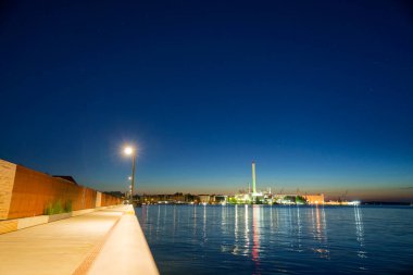 Batı limanı boyunca Flensburg 'da bir yaya bölgesi var. Mavi su akşam ışıklarını yansıtırken RheinMetal fabrikası ve liman kenarları ufkun izini sürüp rıhtımı sakin bir şehir güvertesine çeviriyor..