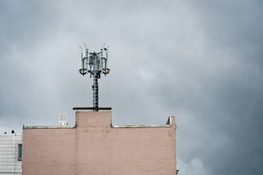 Soluk gül duvarlı eski bir çatı, farklı yönlere yayılmış uzun bir direği ve geniş anten kollarını destekler. Arkasındaki ıslak gri gökyüzü, sahneyi yağmurlu ve yalnız hissettirir..