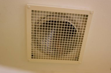 Beyaz bir boru kare bir havalandırma ızgarasının yanındaki duvardan geçiyor. Kurulum, duvarın içinde saklanmak yerine havalandırma ve egzoz akışının görülebildiği basit bir havalandırma sistemine benziyor..