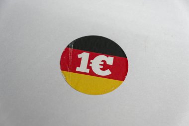 Temiz beyaz bir yüzeyde, üzerinde Alman bayrağı olan bir çıkartma 1 euro yazan cesur bir fiyat notunun yanında duruyor. Etiket, yoldan geçen müşterilerin dikkatini çekmek için ucuz hissettiriyor..