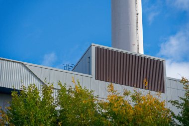 Stadtwerke Flensburg 'un bölgesel ısıtma santrali yüksek baca ve yeşil ağaçların üzerinde temiz tasarım, modern kentsel enerji, sürdürülebilirlik ve yeniliği sembolize ediyor.