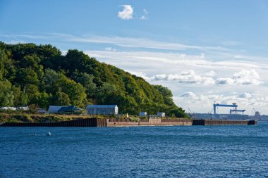 Kiel 'deki sakin bir körfez kıyıda küçük kulübeler ve soluk çadırlar ve etraflarındaki uzun yapraklı ağaçlar gösteriyor. Uzaktaki ağır bitki binaları su hattının üzerinde yükseliyor ve iş sektörüne dair ipuçları veriyor..