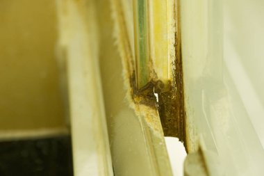 Çürümüş metal bir pencere iskeleti pürüzlü duvarda durur ve çatlamış kaynak onarımlarını ve kahverengi pas katmanlarını gösterir, bütün açıklık zaten defalarca kapatılmış gibi zorlanmış ve yorgun görünür..