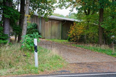 Küçük bir yol, büyük ahşap bir hangarın bulunduğu özel bir avluya doğru tırmanıyor. Karanlık tahtalar ve geniş bir cephe var. Kibar yükseliş ve sessiz eşik, kırsal bölgeyi daha basit ve sakin kılıyor..