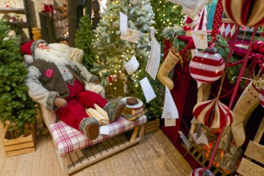 Bir mağaza vitrininde rahat bir sandalyenin yanında küçük yapay ağaçlar var. Orada yumuşak bir Noel Baba bebeği kurdeleler, ışıklar ve çelenkler arasında duruyor. Oyuncak ve ışıltı karışımı sıcak tatil nostaljisi satıyor..