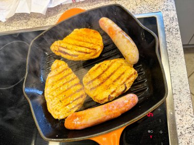 Cızırdayan yağ bir ızgara tavasının sırtlarını işaret ederken, tavuk parçaları ve kısa sosisler birlikte kahverengileşir, akşam yemeği pişerken buhar hafif bir bukle gibi yükselir ve oda sıcak ve lezzetli bir kokuyla dolar..