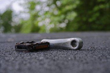 Aşınmış bir bisiklet pedalı yumuşak yeşil bokeh ile kaba asfalta dayanır. Bisiklet temaları, yol güvenliği, tamir kavramları, hareketlilik görselleri ve spor içeriği için ideal.