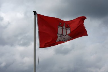 Bulutlu bir gökyüzüne karşı sembolik bir kale amblemiyle kırmızı Hamburg bayrağı. Seyahat temaları, şehir kimliği, bölgesel gurur ve editoryal görseller için ideal.