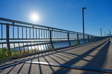 Parlak, berrak bir günde beton zemin ve sakin kıyı sularının üzerinde yan rayları olan uzun bir köprü, yol boyunca uzun sokak lambaları ve güneşte parlayan mavi körfez görünür..
