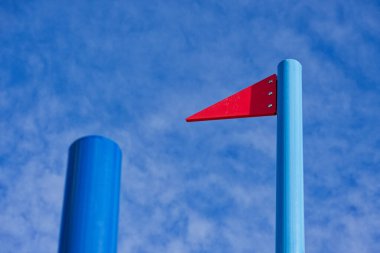 Düz metal bir direk sokağın kenarında durur ve kenarına tutturulmuş sert bir kırmızı üçgen bayrak levhası tutar. Kalın şekli rüzgarsız bir uyarı işareti ya da demirleme rehberi gibi görünür..