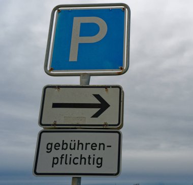 Sessiz caddenin yanında, sağı gösteren beyaz bir okla mavi bir park levhası duruyor. Almanca 'da, sürücülere bu küçük park alanının ödendiğini hatırlatmak için kullanılan kelimeler..