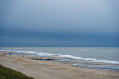 Sylt sahilinin yukarısından mavi deniz geniş kumla karşılaşıyor. Dalgalar ve kumullar arasında yürüyen küçük insanlar. Derin renklerden soluk renklere doğru kayan bir gökyüzü altında..