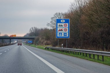 Yol kenarındaki bir panoda hız limiti var. Şehirlerde 50, dışarıda 100, Alman sınırında 130 kilometre. Turuncu bir kamyon gün ışığında otoyoldan geçen bir köprüden geçiyor..