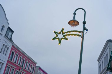 Renkli Alman binaları arasında bir sokak lambası yükseliyor, köknar dallarıyla sarmalanmış parlayan bir yıldız direkten sarkıyor, dar sokağa neşeli bir ışık ekliyor..