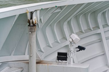 Geminin içinde siyah bir kamera ve küçük bir lamba, metal duvarda bir direği paylaşır, güverte kenarına nişan alır, deniz de yapının ötesine görünür..