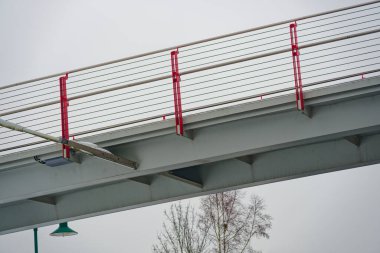 Bulutlu bir kış gününde, çelik kirişli ve kırmızı korkuluklu modern yaya köprüsünün minimalist görüntüsü. Temiz mimari hatlar, endüstriyel tasarım ve kentsel altyapı kavramı.