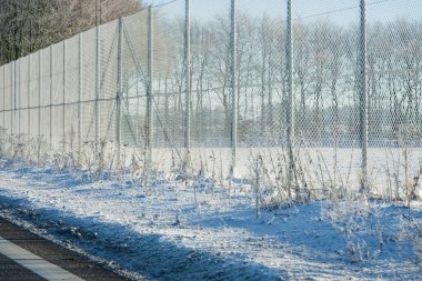Yolun kenarında uzun bir tel örgü var, karla kaplı teller ve direkler, ve kenarlara yayılan pürüzsüz beyaz bir halı, soğuk gün ışığında sahneyi sessizleştiriyor..