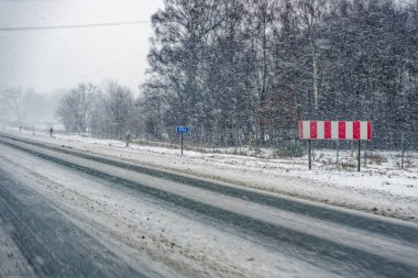 Şiddetli kar yağışında kar ve buzla kaplı, kırmızı ve beyaz uyarı işareti ve yapraksız ağaçlarla kaplı bir A7 E45 yolu..