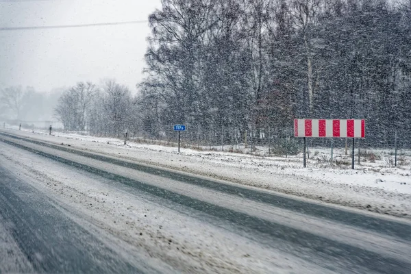 Şiddetli kar yağışında kar ve buzla kaplı, kırmızı ve beyaz uyarı işareti ve yapraksız ağaçlarla kaplı bir A7 E45 yolu..