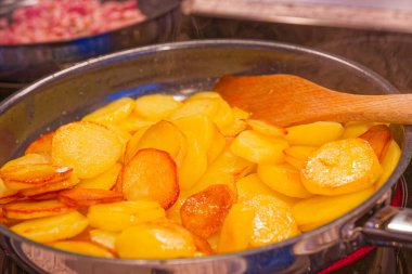 Dilimlenmiş patatesler tavada kızarırken altın çıtır çıtır kenarları ve ahşap bir spatula onları sobanın tepesinde buharla karıştırıyor..