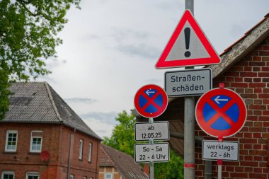 Trafik işaretlerine göre yol hasarı var ve arka planda ağaçlar ve bulutlu gökyüzü olan konut evlerinin yakınında park yasağı yok..