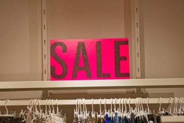 Perakende satış ortamında beyaz askılarda çeşitli giysiler sergilenen büyük siyah harflerle dolu canlı bir pembe satış tabelası..